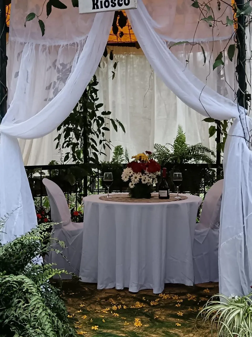 Kiosco decorado para una cena romántica, con mesa para dos, flores y cortinas blancas entre el follaje